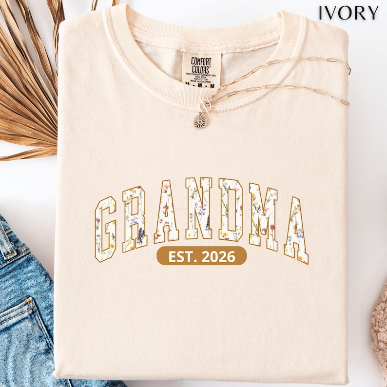 Floral Grandma Est. 2026 shirt with large varsity-style lettering filled with colorful wildflowers, a warm golden outline, and a rounded brown accent beneath the design on a black background.