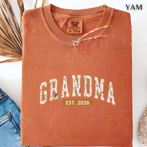 Floral Grandma Est. 2026 shirt with large varsity-style lettering filled with colorful wildflowers, a warm golden outline, and a rounded brown accent beneath the design on a black background.
