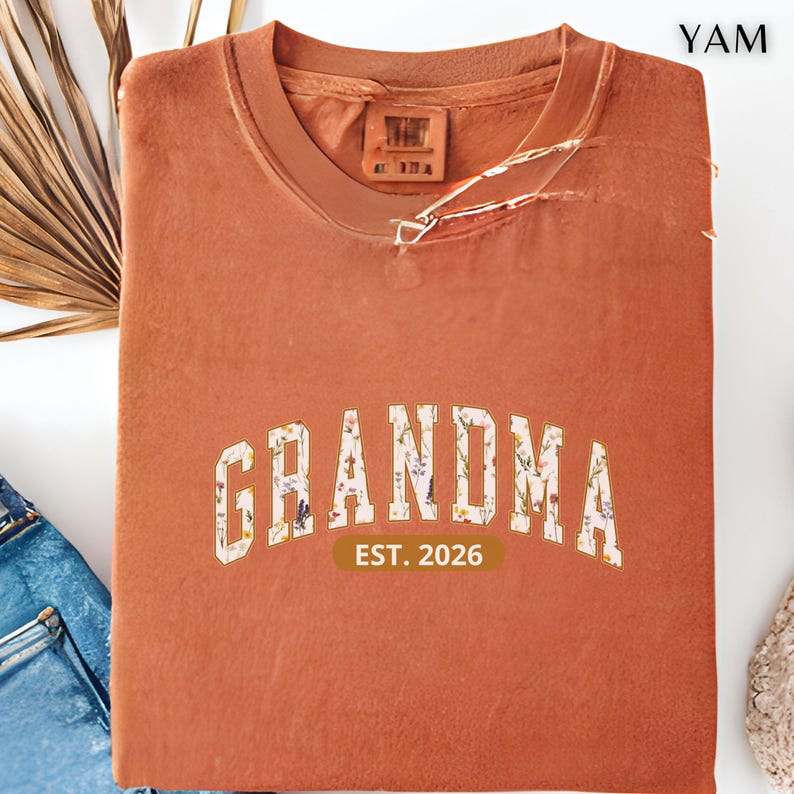 Floral Grandma Est. 2026 shirt with large varsity-style lettering filled with colorful wildflowers, a warm golden outline, and a rounded brown accent beneath the design on a black background.