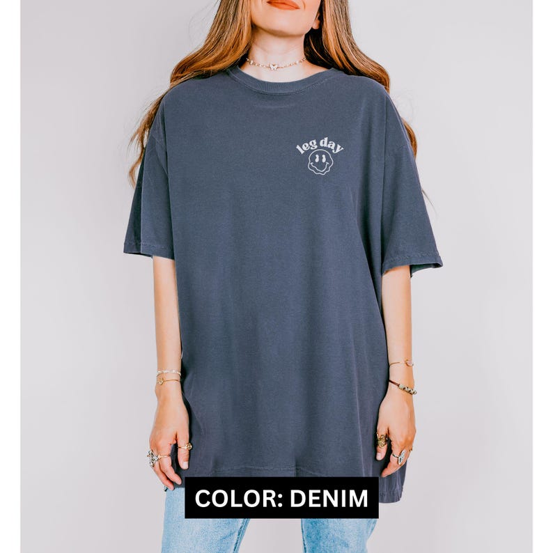 May include: An oversized t-shirt in a denim blue colour with the text "leg day" and a smiley face graphic.