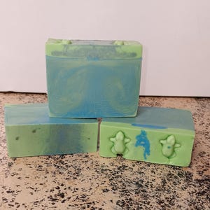May include: Three bars of handmade soap, one with a swirl of green and blue, and two with a green base and blue accents. The two bottom bars have a frog design.