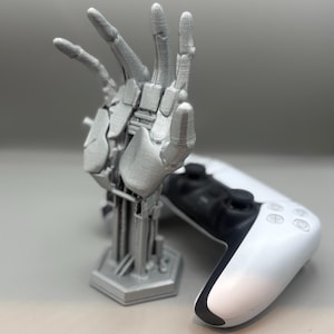 以下が含まれることがあります： 複雑なディテールを持つ銀色のロボットハンドの彫刻。ジェスチャーでポーズをとっています。白と黒のゲームコントローラーが彫刻の隣に置かれており、ゲームをテーマにしていることを示唆しています。彫刻の高さは約15cmです。