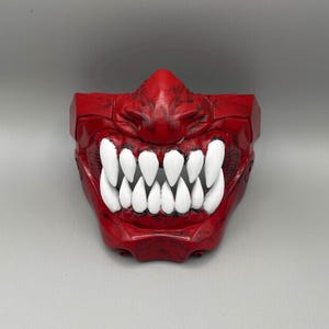 May include: A red Oni mask with prominent white teeth. The mask has a textured finish and a fearsome expression, suitable for cosplay or display. The mask is a striking statement piece.