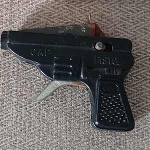 Op de afbeelding: Een zwart speelgoedpistool met een zilveren trekker en de woorden "CAP" en "PISTOL" erop gedrukt. Boven het pistool staat een doosje "BIG BANG CAPS" met de waarschuwing "NIET DICHTERBIJ SCHIETEN".