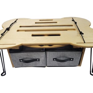 May include: A light-colored wooden lap desk with a unique shape, featuring a slotted top and two gray fabric storage bins below. The desk has black metal hairpin legs and black bungee cords.