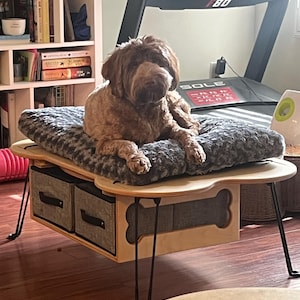 May include: A light brown dog rests on a gray plush dog bed atop a wooden dog bed table. The table has two gray fabric storage drawers and a bone-shaped cutout. Black hairpin legs support the table.