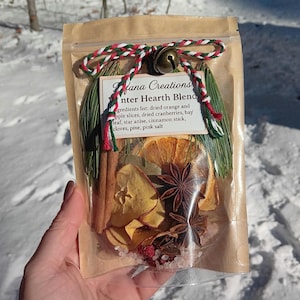 May include: A clear cellophane bag filled with dried orange slices, star anise, cinnamon sticks, and pink salt. The bag is tied with a red, green, and white twine and a small bell. The label reads "fana Creations Winter Hearth Blend."