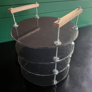 May include: A dark brown, multi-tiered display stand. The stand is constructed with circular, semi-transparent shelves held together by threaded rods and wooden supports. The wooden supports are attached to the top shelf with metal hardware.