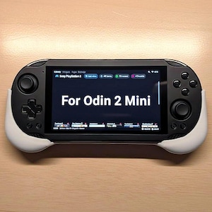May include: A black handheld gaming console with white grips, featuring a screen displaying "For Odin 2 Mini." The console has buttons, a directional pad, and analog sticks. The screen shows game library information.