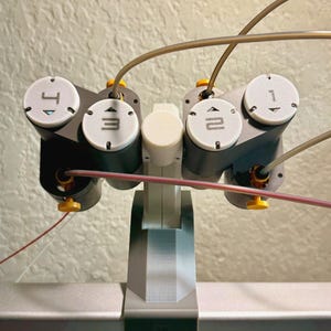 May include: A close-up of a gray and black robotic arm with white circular dials labeled 1, 2, 3, and 4. The arm has yellow and red wires extending from it. The base is gray and white.