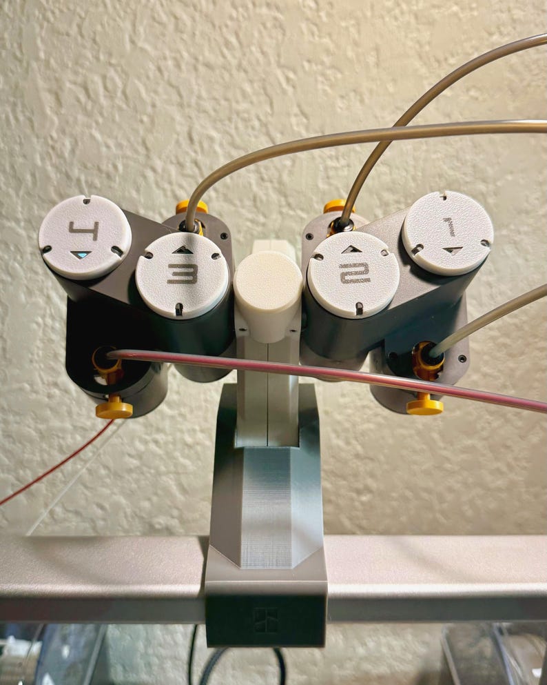 May include: A close-up of a gray and black robotic arm with white circular dials labeled 1, 2, 3, and 4. The arm has yellow and red wires extending from it. The base is gray and white.