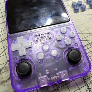 May include: A transparent purple handheld gaming console with a black screen and white buttons. The console features a directional pad, buttons labeled X, Y, A, and B, and two black analog sticks. Additional blue buttons are scattered nearby.