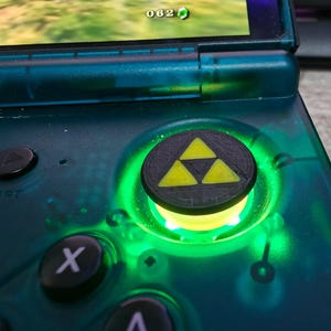 May include: A translucent teal handheld gaming console featuring a circular analog stick cap with a black and yellow Triforce emblem. The area surrounding the analog stick is illuminated by a bright green light, with black buttons and a screen.