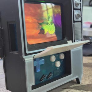 Puede incluir: Un televisor en miniatura de estilo retro con una consola de juegos integrada. La pantalla muestra una escena de dibujos animados. La consola tiene una cruceta azul, botones blancos y las palabras "SELECT" y "START". El televisor es negro con detalles blancos.