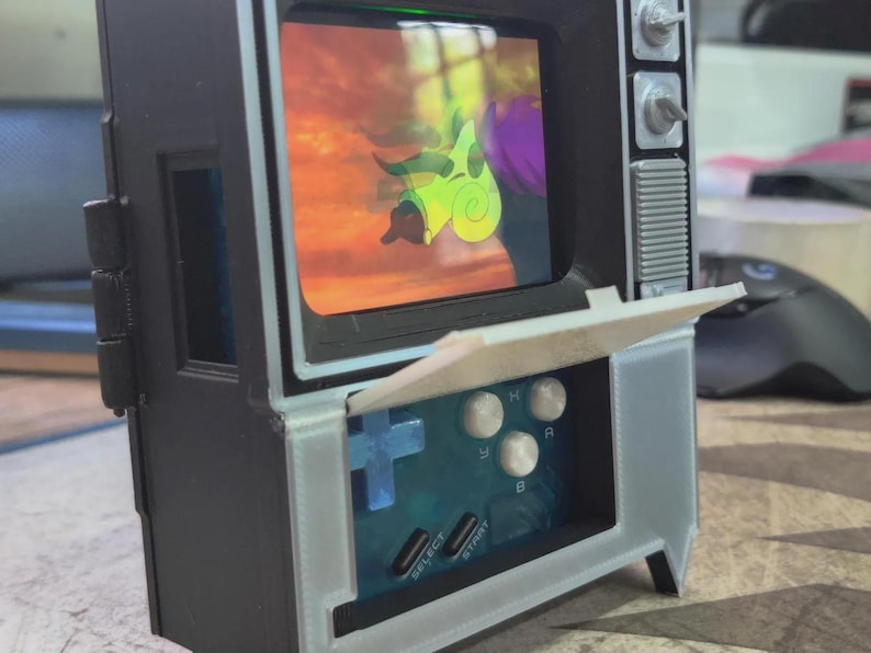 Puede incluir: Un televisor en miniatura de estilo retro con una consola de juegos integrada. La pantalla muestra una escena de dibujos animados. La consola tiene una cruceta azul, botones blancos y las palabras "SELECT" y "START". El televisor es negro con detalles blancos.
