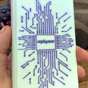 May include: A rectangular, white object with a purple circuit board design. The design features a central square with text and radiating lines. The object is held in a hand.