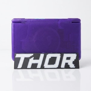 May include: A purple handheld gaming console with a black and white "THOR" logo. The console is closed and has a glossy finish. The logo is rectangular and has a pixelated font.