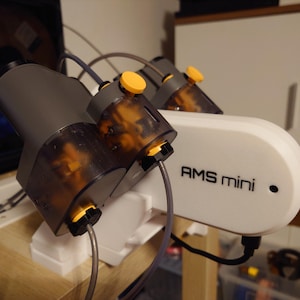 May include: A white and gray AMS mini device with the text "AMS mini" printed on the side. The device has multiple gray and clear components with yellow accents and connected tubes.
