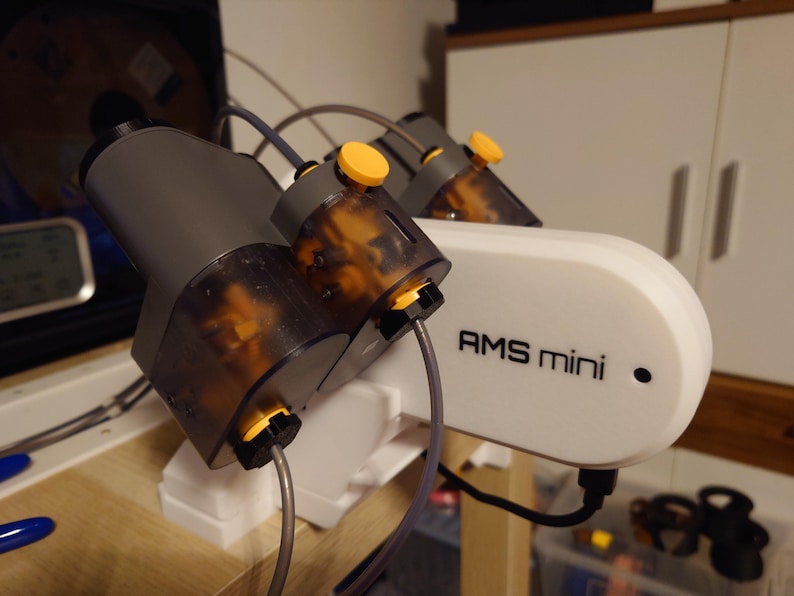May include: A white and gray AMS mini device with the text "AMS mini" printed on the side. The device has multiple gray and clear components with yellow accents and connected tubes.