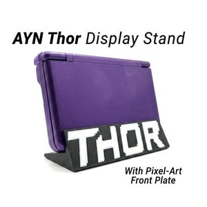 May include: A black display stand with a white pixel-art front plate featuring the text THOR, holding a purple handheld gaming console. The text AYN Thor Display Stand and With Pixel-Art Front Plate is visible in the image.