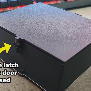 Puede incluir: Una caja negra con tapa texturizada y un cierre seguro para mantener la puerta cerrada. La caja es rectangular y se apoya sobre una superficie plana. El texto "secure latch holds door closed" es visible.