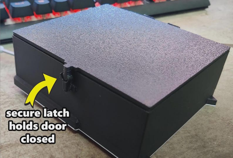 Puede incluir: Una caja negra con tapa texturizada y un cierre seguro para mantener la puerta cerrada. La caja es rectangular y se apoya sobre una superficie plana. El texto "secure latch holds door closed" es visible.