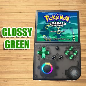 May include: A handheld gaming console with a screen displaying the "Pokémon Emerald Version" game. The console has a transparent black casing with green buttons and a multi-colored light. The words "Glossy Green" are in a green font on the left.