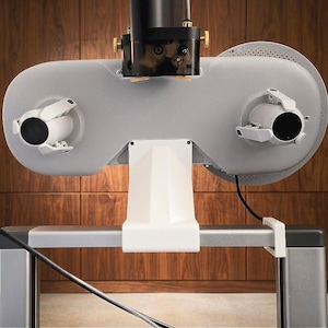 May include: A gray ophthalmic phoropter mounted on a white stand, featuring two circular viewing ports with adjustable white levers, a black central housing with gold adjustment knobs, and a perforated circular disc on the right side.