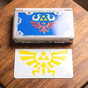 May include: A handheld gaming console featuring a blue and silver top cover with a red and yellow crest design, accompanied by a separate white rectangular decal displaying a yellow winged crest with a central yellow triangle emblem.