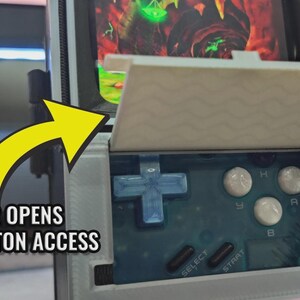 Puede incluir: Una consola de juegos port&aacute;til con un dise&ntilde;o de televisor retro. La pantalla muestra un videojuego, y el dispositivo tiene botones y una cruceta. El texto "TINY DOOR OPENS FOR EASY BUTTON ACCESS" es visible.