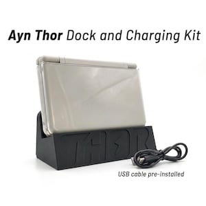 May include: A light gray handheld gaming console rests in a black charging dock with the word THOR embossed on the front. A black USB cable is coiled next to the dock. The text Ayn Thor Dock and Charging Kit appears above, and USB cable pre-installed below.