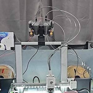 May include: A 3D printer with multiple print heads and clear tubing. The printer has a silver frame and is set up with spools of filament. A box of 3D printing pens is visible in the background.