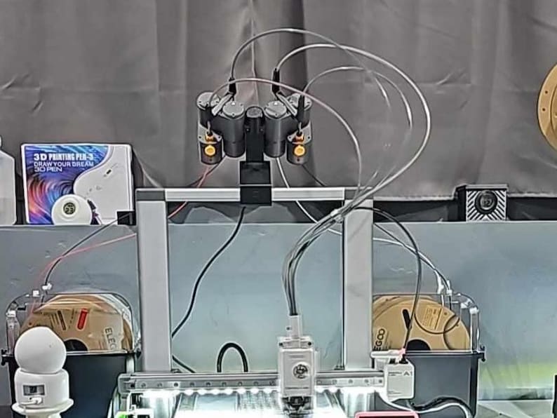 May include: A 3D printer with multiple print heads and clear tubing. The printer has a silver frame and is set up with spools of filament. A box of 3D printing pens is visible in the background.