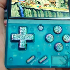 May include: A teal blue handheld gaming console with a silver cross-shaped directional pad, four round buttons, and two rectangular buttons labeled "SELECT" and "START".