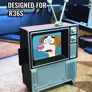 Puede incluir: Un televisor en miniatura de estilo retro con un perro de dibujos animados en la pantalla. El televisor es negro con un panel frontal azul claro y una parte superior rosa. El texto "DESIGNED FOR R36S" est&aacute; encima del televisor.