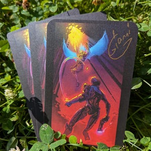 May include: A rectangular trading card with a black border featuring a colourful illustration of a robotic figure in red armour facing a winged character with blue wings and a golden helmet. The card includes a gold signature reading Gianni.