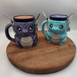 May include: Two dragon-shaped mugs with lids and silver spoons. One mug is dark purple with a light gray belly, and the other is light blue with a light gray belly. Both feature large eyes, textured scales, and a handle shaped like a tail.