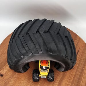 Puede incluir: Un neum&aacute;tico negro grande se arquea sobre un peque&ntilde;o juguete de monster truck amarillo con llamas rojas y naranjas. El juguete tiene neum&aacute;ticos negros y est&aacute; colocado sobre una superficie de madera.