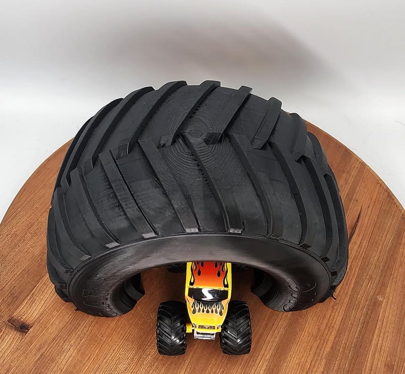 Puede incluir: Un neum&aacute;tico negro grande se arquea sobre un peque&ntilde;o juguete de monster truck amarillo con llamas rojas y naranjas. El juguete tiene neum&aacute;ticos negros y est&aacute; colocado sobre una superficie de madera.