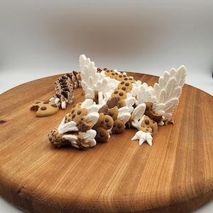 May include: A decorative sculpture of a dragon, featuring a segmented body and wings. The dragon is primarily brown with dark brown spots and white accents. It is displayed on a round wooden surface.