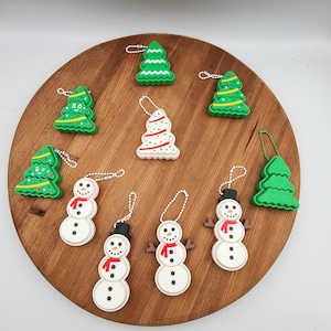 May include: A collection of Christmas ornaments, including green Christmas trees and snowmen. The snowmen have black top hats, red scarves, and stick arms. The ornaments are on a wooden surface.