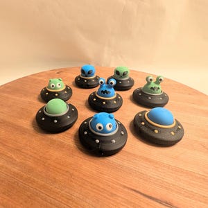 May include: Eight small, black flying saucer toys with different colored alien figures inside. The aliens have green, blue, and light blue skin tones with various facial expressions.