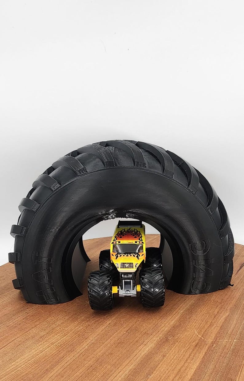 Puede incluir: Un juguete de cami&oacute;n monstruo amarillo y negro est&aacute; posicionado debajo de un gran arco de llanta negro. El cami&oacute;n de juguete tiene llantas negras grandes y un dise&ntilde;o de llama. El arco est&aacute; hecho de un material negro y se asienta sobre una superficie de madera.