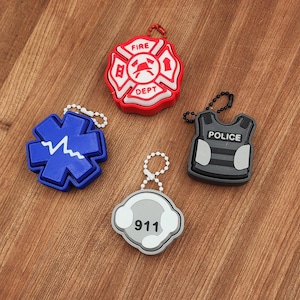 May include: Four keychains on a wooden surface. The keychains include a red and white fire department emblem, a blue medical symbol, a gray police vest, and a gray 911 emblem. Each keychain has a metal chain or ball chain.