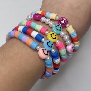 May include: A collection of colorful beaded bracelets with smiley face charms and faux pearl accents. The bracelets feature a variety of colors, including pink, blue, yellow, and orange. The bracelets are stacked on top of each other.