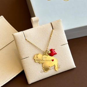 Op de afbeelding: Goudkleurige ketting met een hanger in de vorm van Venezuela, met de inscriptie "VENEZUELA". Een kleine rode hartvormige bedel is bevestigd. De ketting wordt gepresenteerd op een beige kaart, met een witte doos op de achtergrond.