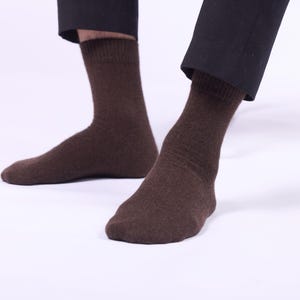 May include: A pair of brown socks, visible from the ankles down, against a white background. The socks are a solid, dark brown color and appear to be made of a soft, textured material. The legs are partially visible, wearing black pants.