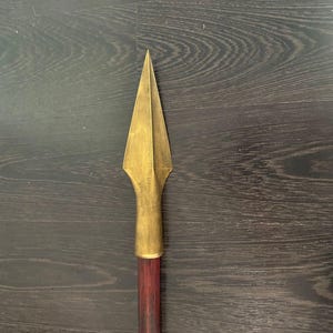 Puede incluir: Una punta de lanza dorada con un mango de madera. La punta de lanza es puntiaguda y tiene un diseño ligeramente curvo. El mango es de color marrón oscuro y parece estar hecho de madera.