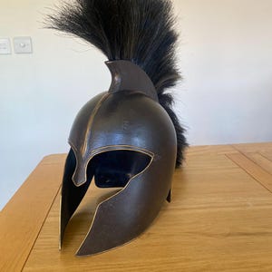 Puede incluir: Un casco espartano marrón oscuro con una pluma de crin de caballo negra. El casco tiene un ribete dorado y una cresta elevada.
