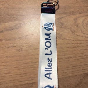 Pode incluir: Um cord&atilde;o branco com o texto "Allez L'OM" em azul, juntamente com o log&oacute;tipo da equipa. O cord&atilde;o tem um fecho met&aacute;lico e um porta-chaves. &Eacute; provavelmente um acess&oacute;rio de f&atilde;.